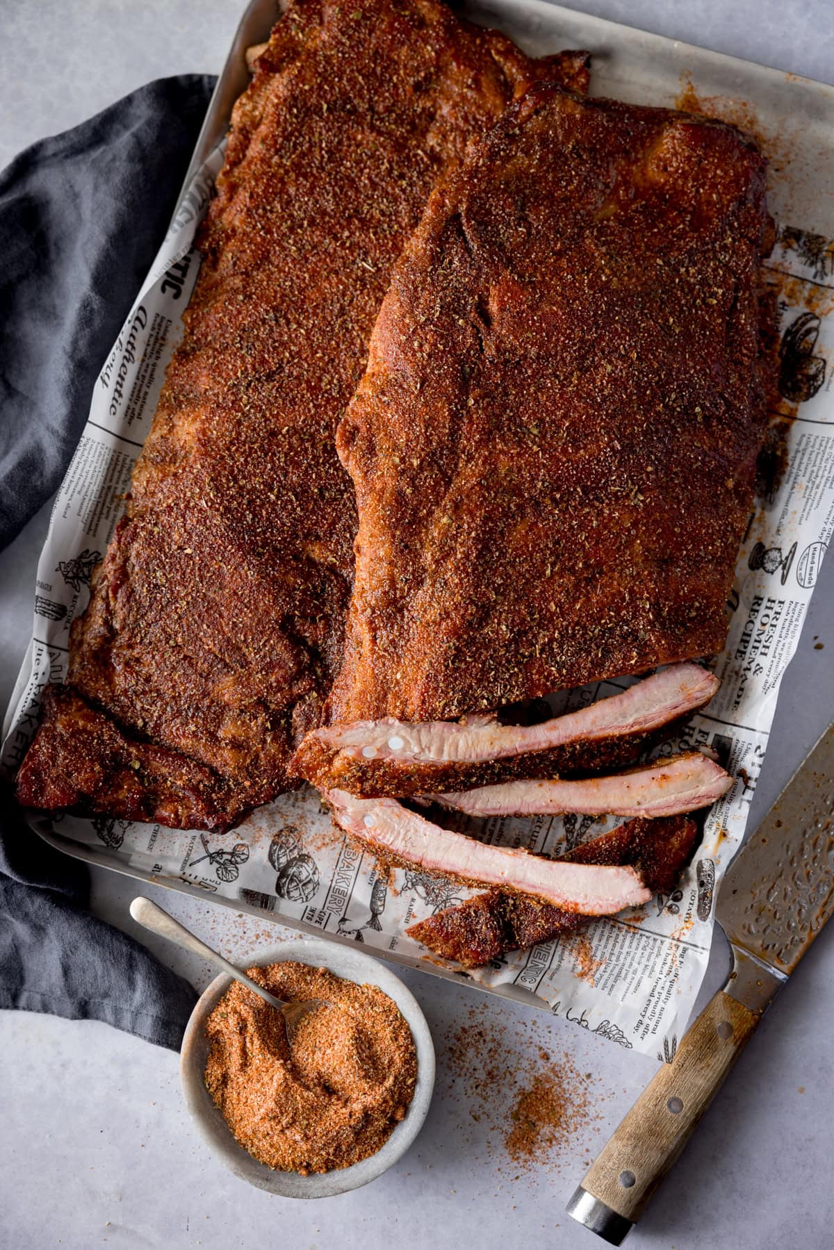 A tall, overhead image of dry rub BBQ ribs, there are two racks of the ribs laid out on a silver tray that is lined with newspaper. The central rack of ribs is cut into and the three of the ribs are separated so you can see the ribs. On the bottom left of the background, you can see a slate grey napkin tucked underneath the tray, in the bottom centre there is the dry rub seasoning in a light grey bowl with a silver teaspoon sticking out of it. On the bottom right of the image, there is a large silver knife with a wooden handle set at an angle. This is all set on a light grey background.