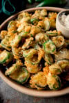 A tall image of a light brown bowl of fried Jalapenos. In the top right of the bowl, there is a light grey dish nestled in the fried Jalapenos, which has garlic mayo in it. In the top left of the background, you can slightly see a dark green napkin. This is all set on a grey surface.