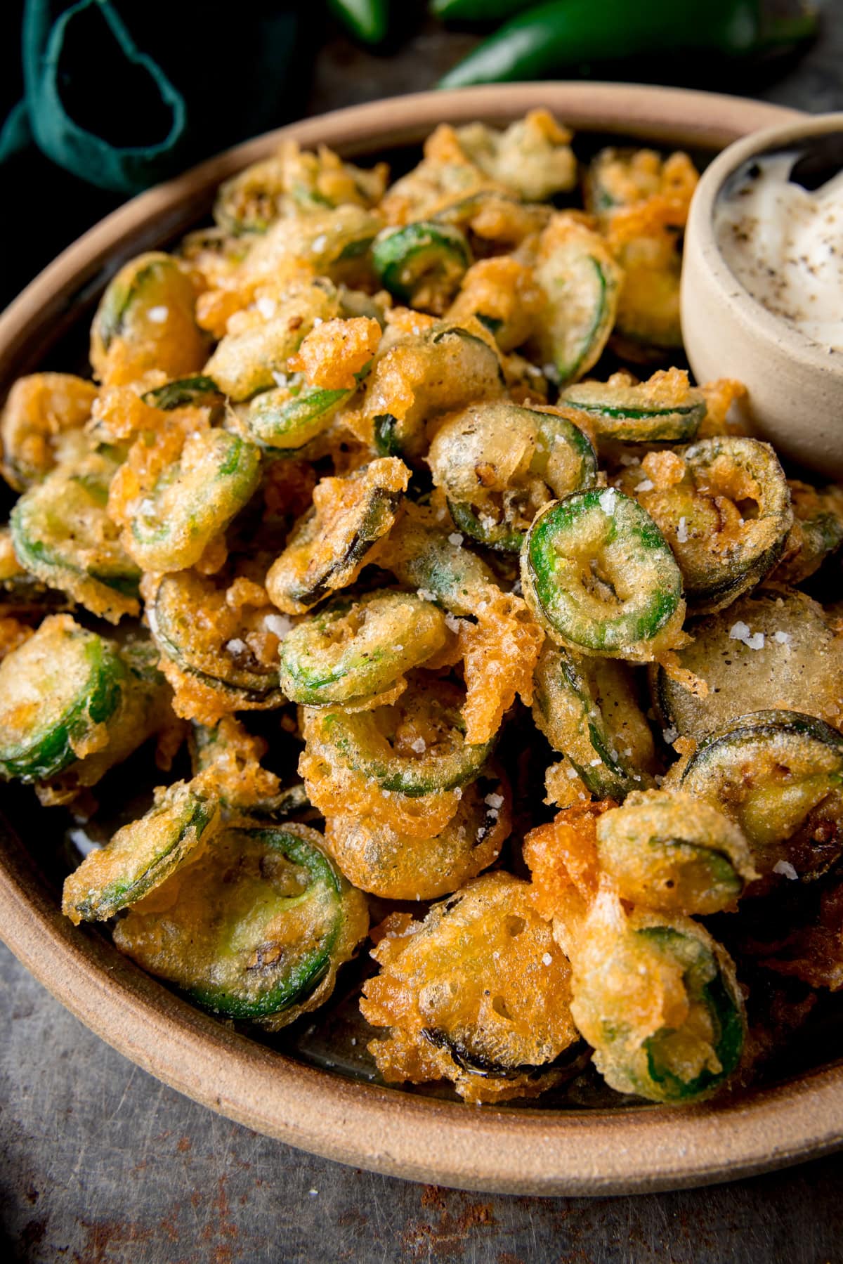 A tall image of a light brown bowl of fried Jalapenos. In the top right of the bowl, there is a light grey dish nestled in the fried Jalapenos, which has garlic mayo in it. In the top left of the background, you can slightly see a dark green napkin. This is all set on a grey surface.