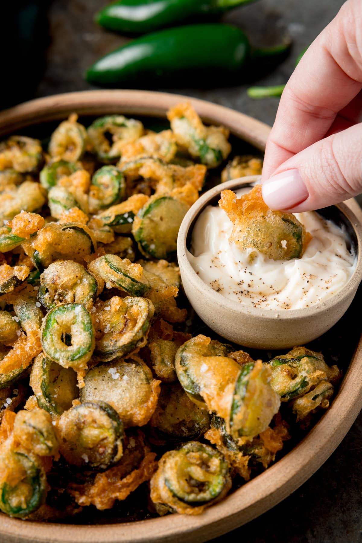 A tall image of a light brown bowl of fried Jalapenos. In the top right of the bowl, there is a light grey dish nestled in the fried Jalapenos, which has garlic mayo in it. There is a hand coming from the right of the screen, which is holding a fried Jalapeno slice and dipping it in the dish of garlic mayo. In the top middle of the background, you can see two fresh, green Jalapenos next to each other. This is all set on a grey surface.