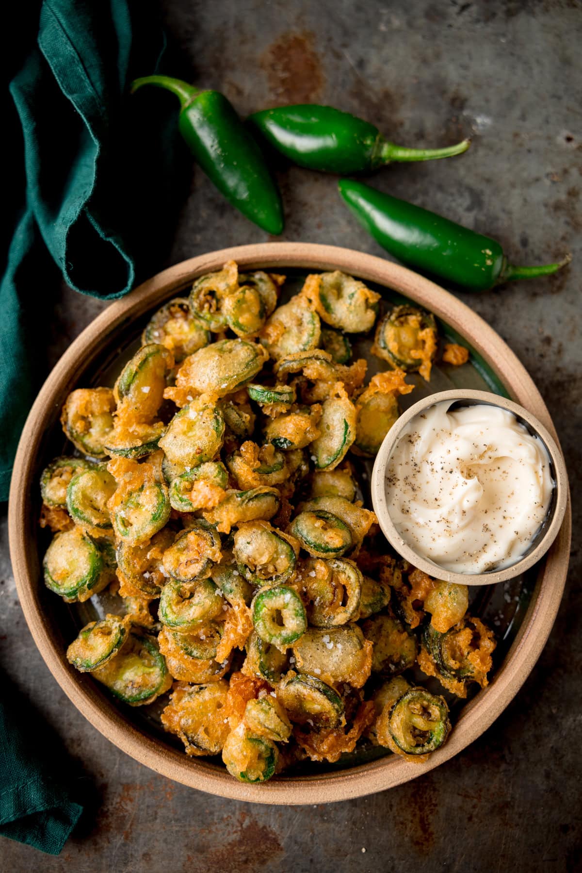 A tall, overhead image of a light brown bowl of fried Jalapenos. In the top right of the bowl, there is a light grey dish nestled in the fried Jalapenos, which has garlic mayo in it. In the top left of the background, there is a dark green napkin. In the top middle of the background, you can see three fresh, green Jalapenos next to each other. This is all set on a grey surface.