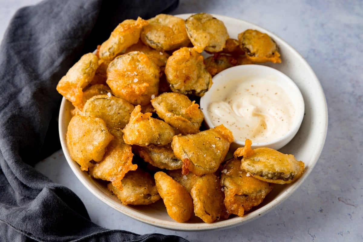 A bowl of crispy battered fried pickles with a small bowl of garlic mayonnaise.
