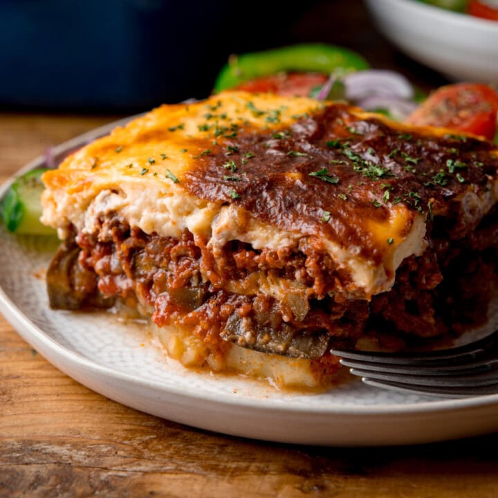 A square, close-up shot of a serving of Moussaka, on a white plate. On the right, you can slightly see a silver fork on the plate with the moussaka. In the background, you can also see some salad on the plate behind the moussaka. You can also see a white bowl, also filled with salad on the navy blue background. This is all set on a wooden surface.
