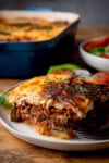 A tall, close-up shot of a serving of Moussaka, on a white plate. On the right, you can slightly see a silver fork resting on the plate in front of the moussaka. In the background, you can also see some salad on the plate behind the moussaka. You can also see a white bowl, also filled with salad on the right of the background. On the left of the background, you can see a navy blue dish, containing the rest of the moussaka. This is all set on a wooden surface, with a slate grey background.