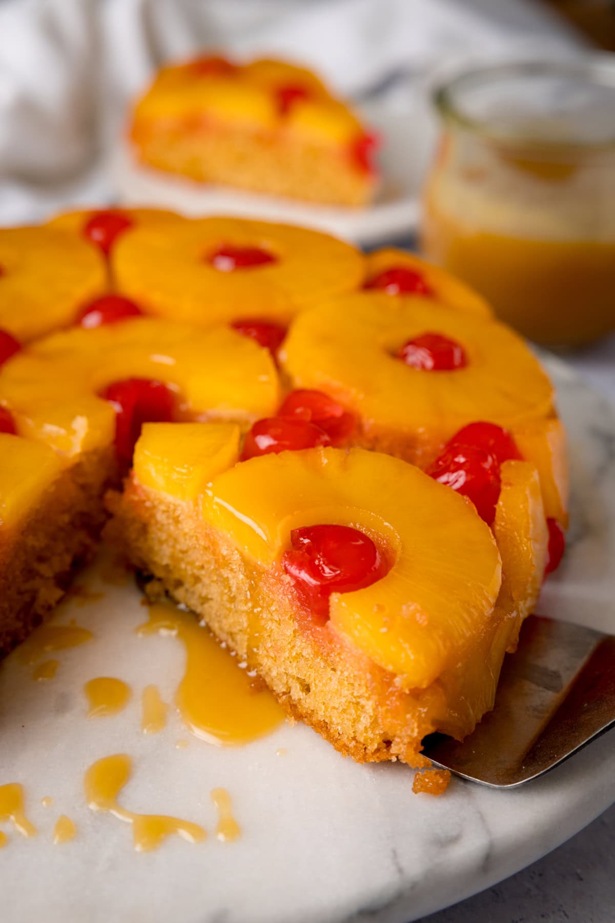 A tall shot of pineapple upside-down cake. In the centre foreground, there is the whole pineapple upside-down cake, with a slice missing. The cake is on a white marble cake stand. In the background, there is a slice of cake on a white plate, with a white napkin tucked behind it. To the right of the background, there is a clear jar of pineapple syrup. This is all set on a white surface.