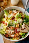 A tall, overhead shot of chicken Caesar salad. The chicken salad is in a white bowl, in the centre of the image. There are the handles of two silver forks sticking out of the salad. The bowl is on top of a wooden board. In the top left of the image, on top of the wooden board, there is a small pile of crispy croutons and a small glass jar of Caesar dressing. In the bottom right of the image, there is a navy blue napkin, tucked around the wooden board. This is all set on a white surface.