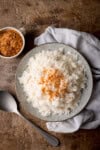 A tall, overhead shot of Stovetop Coconut Rice. The rice is on a light grey plate in the centre of the image. To the right of the plate, there is a light grey napkin tucked beneath the plate. To the bottom left of the plate, there is a silver spoon resting on the surface at an angle, and to the top left of the plate, there is a white ceramic mug filled with toasted shredded coconut flakes. This is all set on a wooden surface.