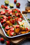 A tall shot of grilled fruit salad in a white tray with a blue lining. There is a dollop of Greek yoghurt on the bottom right side of the tray. The tray is on top of a wooden cutting board, set at an angle. There are strawberries scattered around the wooden board and the surface. In the top right corner of the image, you can see a small glass bowl filled with honey, and in front of it, in the foreground, a honey stirrer, covered in honey which is being drizzled onto the fruit. Also, at the top of the image, you can see a few lime wedges, on the surface. This is all set on a light grey background.