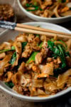 A tall shot of Pad See Ew served in a white bowl. In the centre of the image, on the top rim of the main bowl, there is a pair of wooden chopsticks with a blue handle resting on the bowl. In the top right of the background, you can see a second white bowl of Pad See Ew. Next to it, in the top left corner of the background, there is a white dish filled with dried chilli flakes. This is all set on a wooden background.