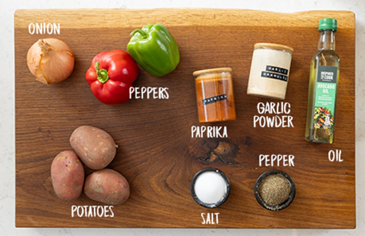 A wide, overhead shot of the ingredients for Air Fryer Breakfast Potatoes. The ingredients are laid out on a wooden cutting board, which is on a white marble surface. The ingredients are labelled in white text below them. They are as follows: onion, peppers, potatoes, salt, pepper, paprika, garlic powder, and oil.