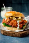 A tall image of a Buffalo Chicken Burger is placed on top of a square of black and white newspaper parchment, which is on top of a dark wooden cutting board. In the left of the background, you can see a clear glass jar of homemade ranch, with a silver spoon sticking out of it. This is all set on a navy blue background.