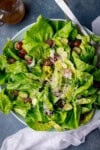 A tall, overhead shot of Butter Lettuce Salad in a large white bowl. The bowl with the salad is placed in the centre of the image, in the top right corner of the bowl there are two silver metal salad salad servers sticking out of the dish. In the top left corner of the image, you can see a clear glass jug with the rest of the salad dressing in it. Just tucked below the bottom right-hand side of the bowl, there is a white napkin. This is all set on a light blue/grey background.