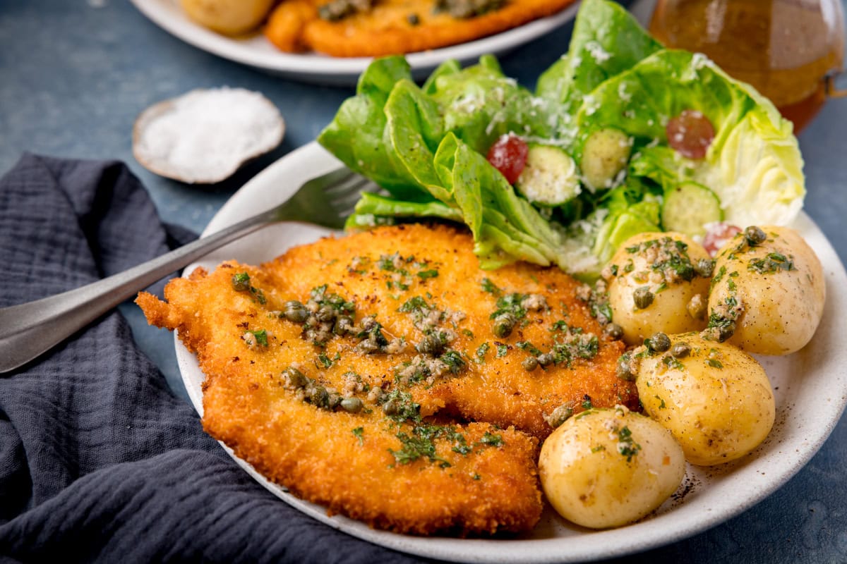 Flat breaded crispy chicken topped with caper butter on a white plate with boiled potatoes and salad