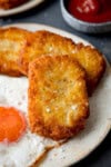 A tall, close-up shot of three hash browns on the side of a large white plate, which has a fried egg in the centre. In the top left of the image, you can slightly see a second white plate. In the top right-hand side of the image, there is a dark blue dish filled with ketchup. This is all set on a navy blue background.