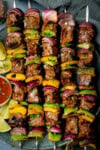 A tall overhead shot of steak kebabs. There are five kebabs placed on a blue plate, beside the skewers, on the left side of the plate there is a small white dish with chilli sauce and four wedges of lime. In the top left of the image, you can slightly see a clear glass jar with dried chilli flakes in it and scattered around it. In the top right of the image, there is a navy blue napkin tucked beneath the main plate. There are dried chilli flakes and shredded coriander scattered around the surface. This is all set on a light blue/grey surface.