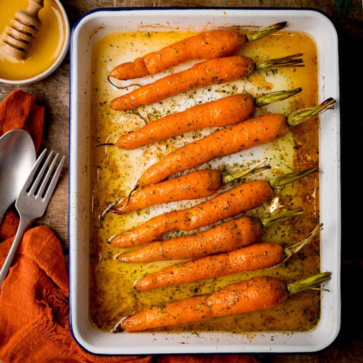 A square, overhead shot of Honey Roasted Carrots in a white tray. To the top left of the tray, there is a small white dish filled with honey, with a wooden honey stirrer in it. Below that, there is an orange napkin with a silver fork and spoon placed on top of it at an angle. This is all set on a wooden surface.