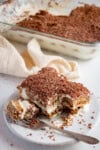 A close up image of a stone plate with a slice of tiramisu sat on top. There's a fork on the plate that has taken a bite out of the tiramisu. The plate is sat on a light coloured board with a glass dish with the rest of the tiramisu in the background slightly out of focus.