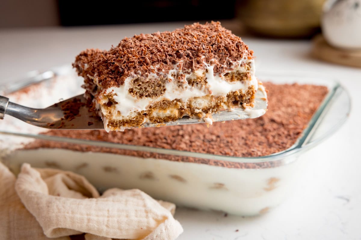 A slice of tiramisu taken out of rectangular dish and raised above on a cake server.