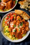 A tall, overhead shot of slow cooker chicken curry on a bed of pilau rice, with some tomato and onion salad, and a corner of naan all in a small white bowl. There is also a silver spoon sticking out of the left-hand side of the dish. In the top left corner of the image, you can slightly see the corner of another bowl of curry. In the top right corner of the image, you can see some torn-up naan on the surface and directly below the main bowl of curry, there is a navy blue napkin tucked beneath the bowl. This is all set on a dark blue background.