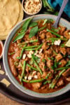 A tall, overhead shot of Slow Cooker Thai Red Beef Curry in a light blue bowl. There is a silver spoon sticking out of the dish. To the left of the bowl, you can slightly see a small white dish filled with sweet chilli sauce, above that you can see a stack of popadoms. Directly above the main bowl, you can see a small white dish filled with peanuts. In the top right corner of the image, there is a blue napkin tucked beside the bowl. All of this is on top of a black tray, which is on top of a wooden surface.