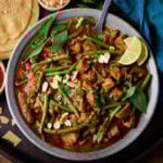 A square, overhead shot of Slow Cooker Thai Red Beef Curry in a light blue bowl. There is a silver spoon sticking out of the dish. To the left of the bowl, you can slightly see a small white dish filled with sweet chilli sauce, above that you can see a stack of popadoms. Directly above the main bowl, you can see a small white dish filled with peanuts. In the top right corner of the image, there is a blue napkin tucked beside the bowl. All of this is on top of a black tray, which is on top of a wooden surface.