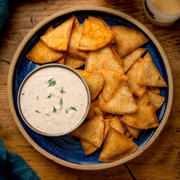 A square, overhead shot of baked tortilla chips on a blue plate with a light brown rim. There is a small dish of Buffalo Ranch Dressing also on the dish. In the top right corner of the image, there is a small glass jar of garlic powder, and in the bottom left corner of the image, you can slightly see a teal napkin. This is all set on a wooden cutting board.