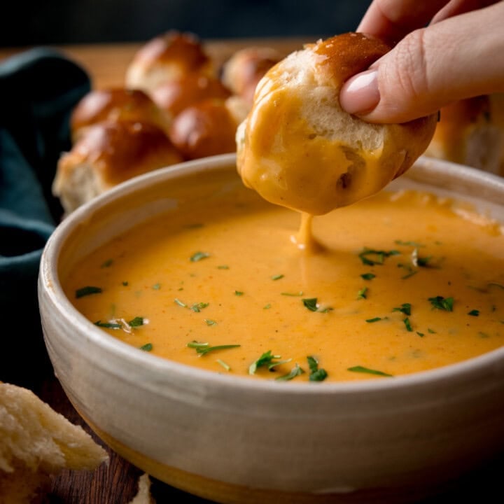 A hand dips a piece of bread into a bowl of creamy orange beer cheese dip, garnished with chopped herbs. More bread rolls are visible in the background on a wooden surface.