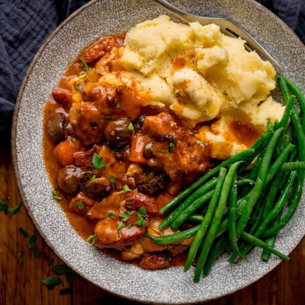 A square, overhead shot of Chicken Chasseur served in a light grey bowl with mashed potatoes and green beans. There's a silver fork resting on the plate. In the top left corner of the image, there is a dark blue napkin tucked beneath the plate. This is all set on a wooden surface.