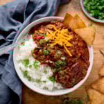A bowl of chilli con carne with kidney beans, ground beef, cheese, and cilantro over white rice. Tortilla chips on the side.