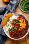 A bowl of chilli con carne with kidney beans, minced beef, cheese, and coriander over white rice. Tortilla chips on the side and a blue napkin to the side of the bowl.
