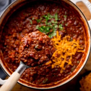 A ladle lifts a hearty chili from a pot, topped with cilantro and shredded cheese, with bowls of herbs and cheese nearby.