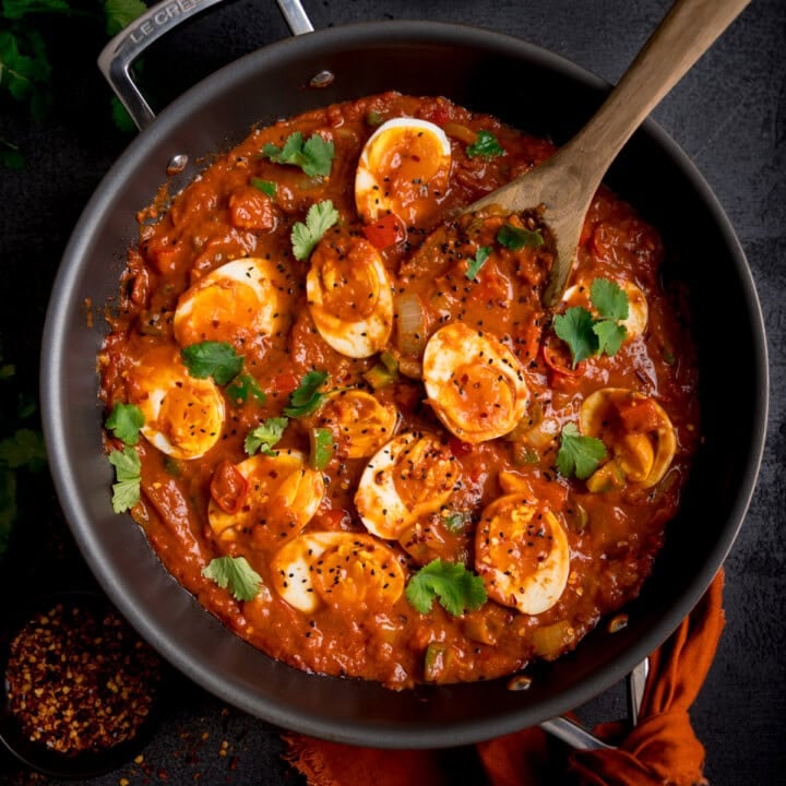 A frying pan filled with spicy egg curry, part of a delightful egg curry recipe, is topped with halved boiled eggs and garnished with fresh coriander. A wooden spoon rests in the curry, while a nearby small bowl of red chilli flakes sits on the dark surface.