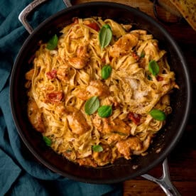 A square, overhead shot of Marry Me Chicken Pasta in a black pan. To the left of the pan, there is a teal napkin tucked beside it. This is all set on a wooden surface.