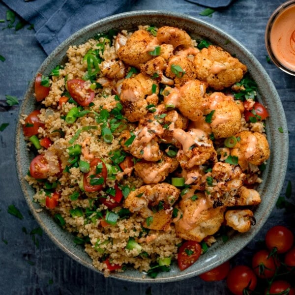 A square, overhead shot of Smoky Cauliflower Salad in a large blue bowl. In the bottom right corner, you can see a small pile of cherry tomatoes, and in the top right corner, you can see a small glass jar of siracha mayo. This is all set on a light blue surface.