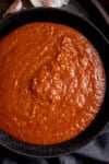 A rich coconut curry sauce in a black bowl on a dark surface, accompanied by hints of garlic cloves and a cloth in the background.