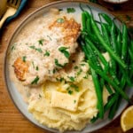 A plate with garlic parmesan chicken in creamy sauce, mashed potatoes with a knob of butter, and green beans, garnished with parsley.