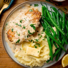 A plate with garlic parmesan chicken in creamy sauce, mashed potatoes with a knob of butter, and green beans, garnished with parsley.