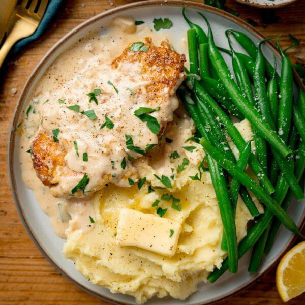 A plate with garlic parmesan chicken in creamy sauce, mashed potatoes with a knob of butter, and green beans, garnished with parsley.