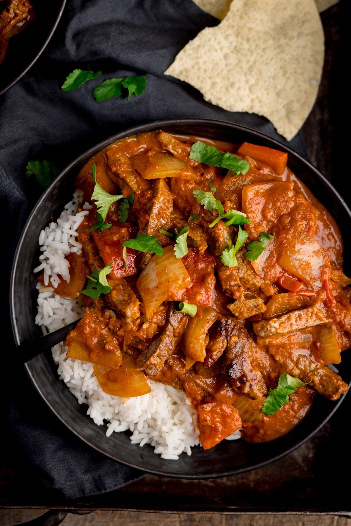 Overhead image of a bowl of curry and rice garnished with coriander on a dark tray. There are some broken up poppadoms in the background.