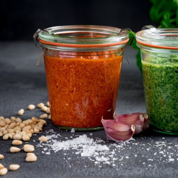 Two glass jars, one with roasted red pepper pesto and another with green pesto, surrounded by pine nuts, garlic cloves, and coarse salt on a dark surface.
