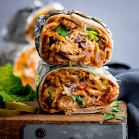 Two halves of a rotisserie chicken burrito packed with rice, rotisserie chicken, cheese, beans, and vegetables are wrapped in foil and garnished with coriander. They sit stacked on a wooden surface.