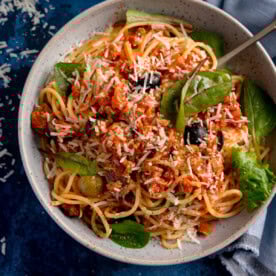A bowl of tuna spaghetti with tomato sauce, black olives, and leafy greens, topped with grated cheese.