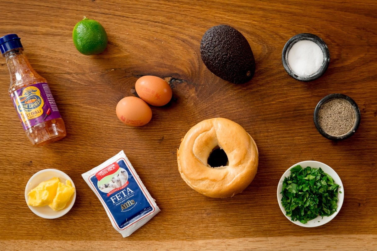 Ingredients for avocado egg bagel on a wooden board. Including: a bagel, eggs, avocado, a lime, feta cheese, coriander (cilantro), butter, sweet chilli sauce, salt and pepper.