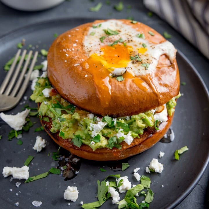 A bagel with a fried egg, smashed avocado, crumbled cheese, and chopped herbs sits on a dark plate with a fork beside it.