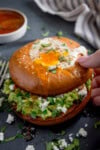 A hand lifts a bagel topped with a fried egg, revealing creamy avocado, feta cheese, and fresh herbs inside.