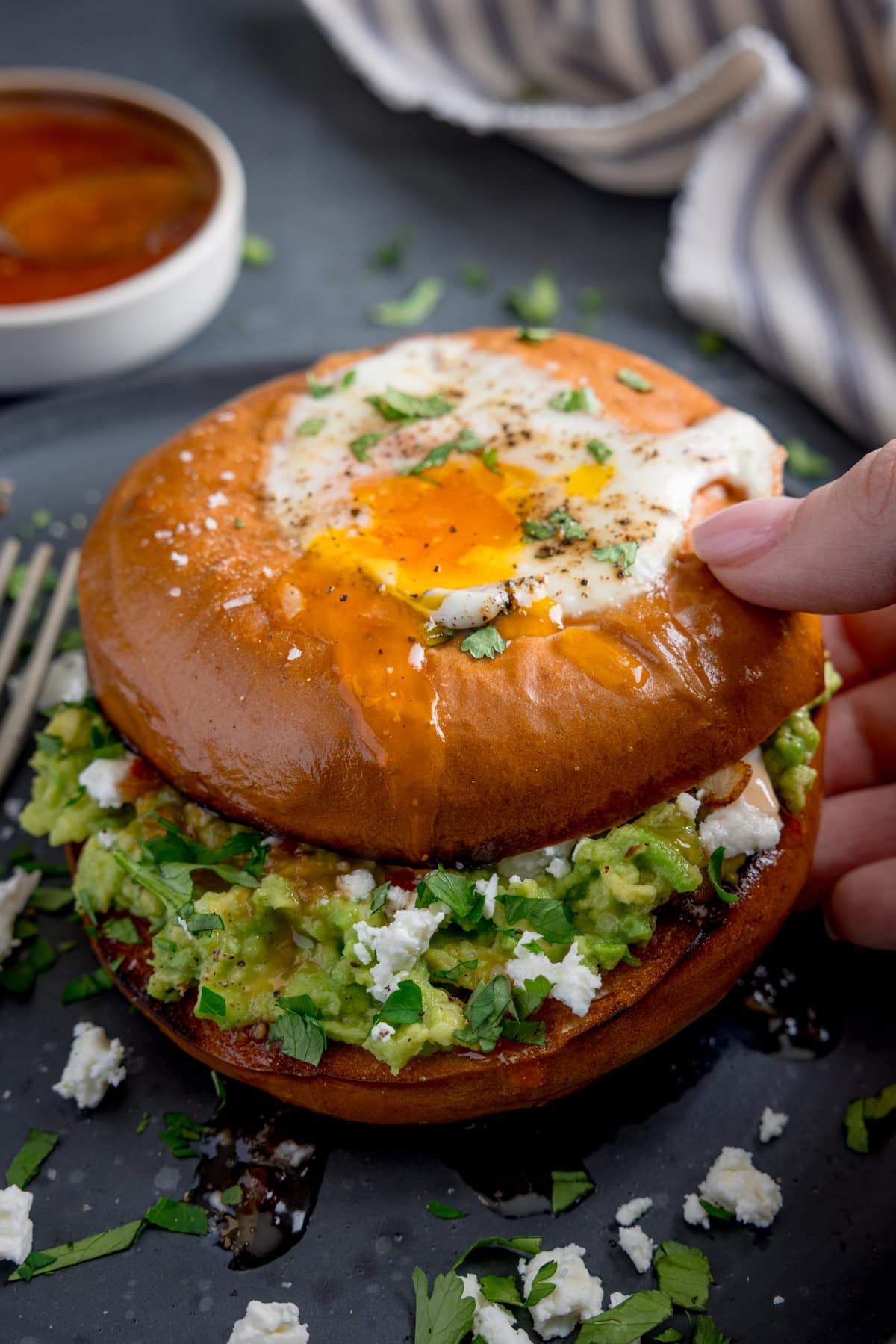 A hand lifts a bagel topped with a fried egg, revealing creamy avocado, feta cheese, and fresh herbs inside.