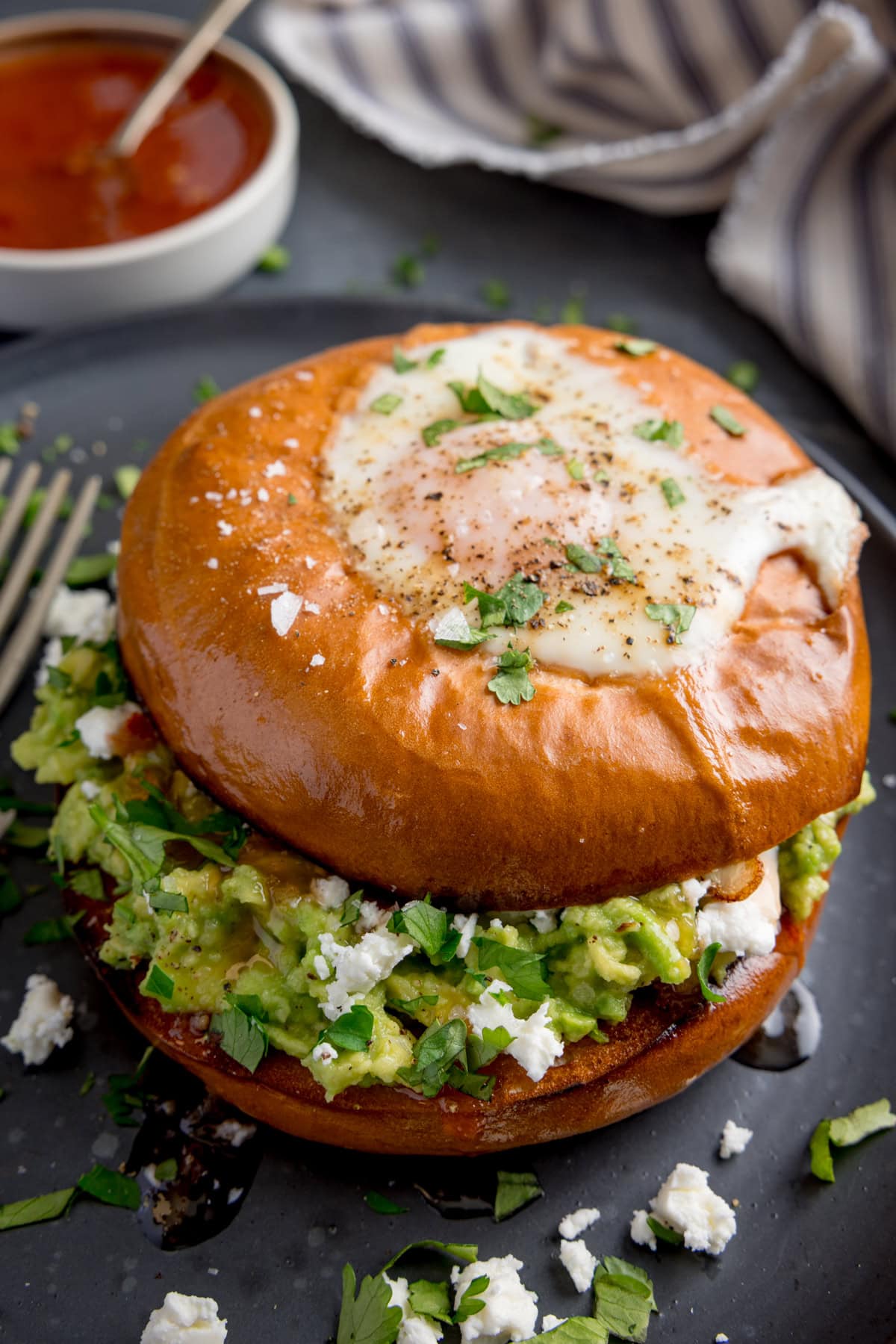 A bagel with a fried egg, smashed avocado, crumbled cheese, and chopped herbs sits on a dark plate with a fork beside it.