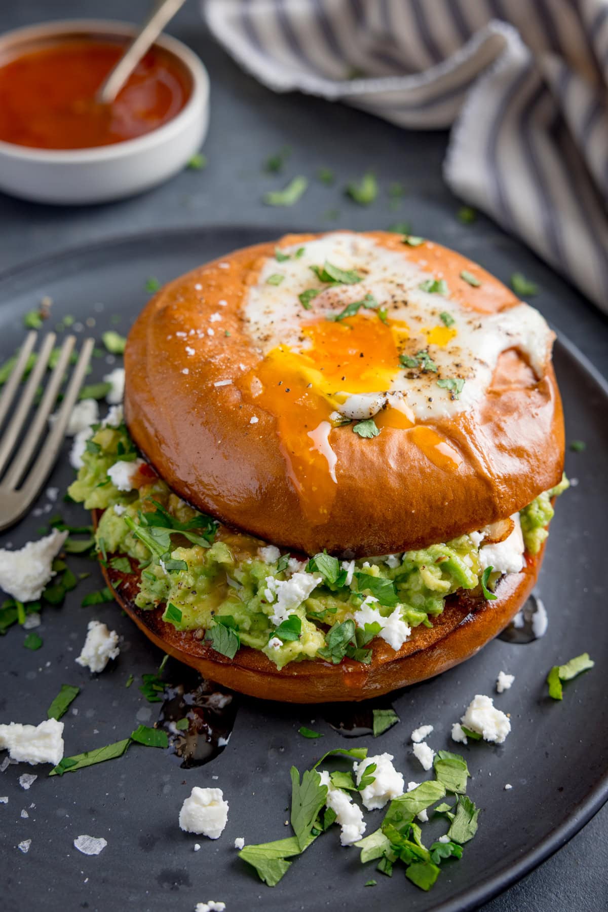 A bagel with a fried egg, smashed avocado, crumbled cheese, and chopped herbs sits on a dark plate with a fork beside it.