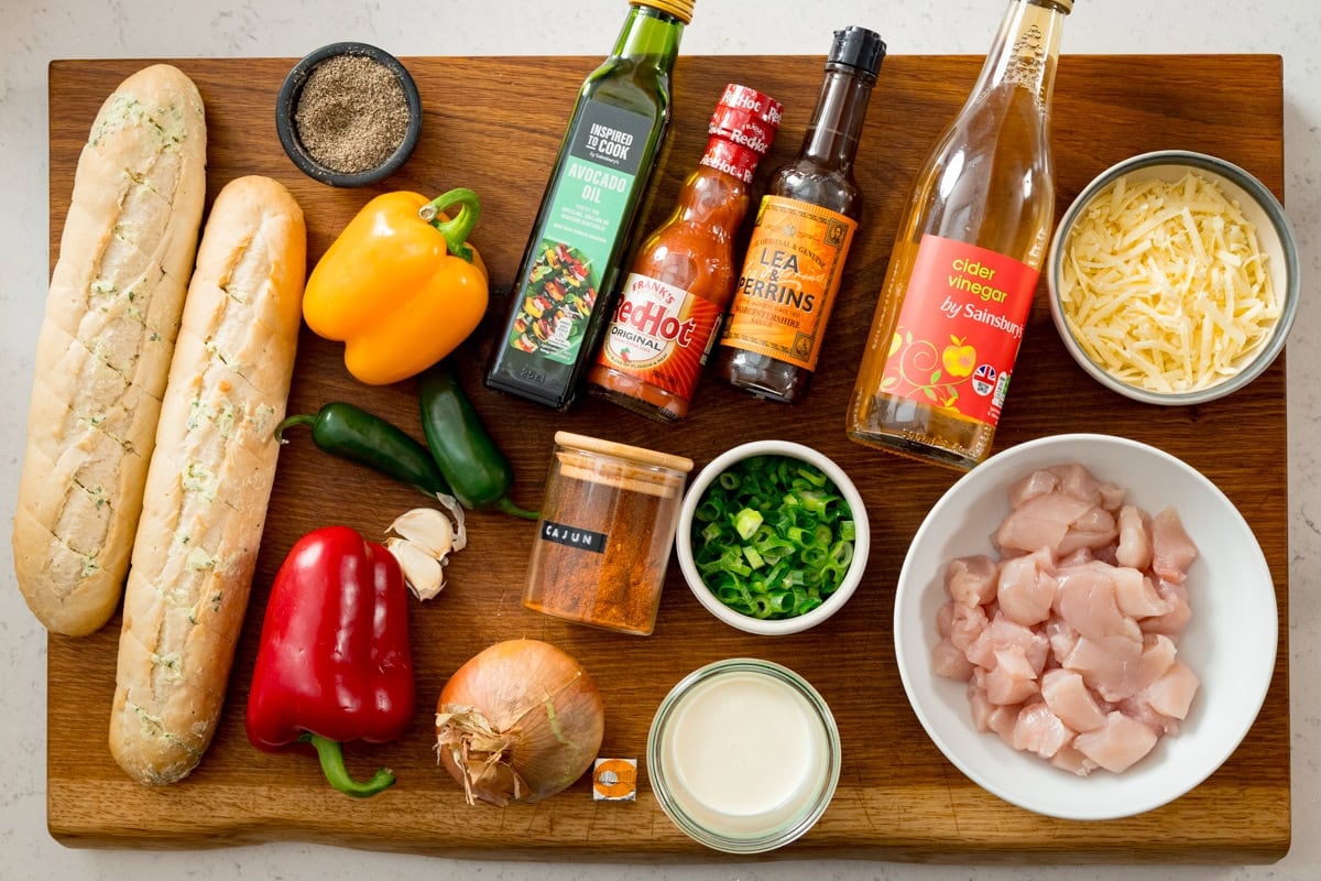 Ingredients for Buffalo Chicken Garlic Bread on a wooden board - including 2 sliced garlic baguettes, peppers, jalapeños, garlic, onion, spring onions, chicken pieces, cheese, seasoning, sauces, cider vinegar, avocado oil and cream.