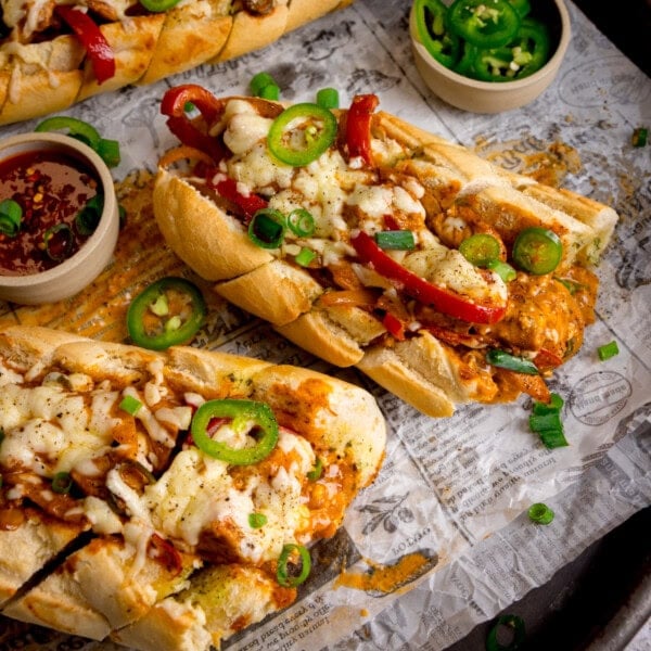 Toasted garlic bread filled with buffalo chicken, melted cheese, sliced red peppers, and topped with jalapeño slices and spring onions, served on baking parchment with a side of sauce and extra jalapeños.